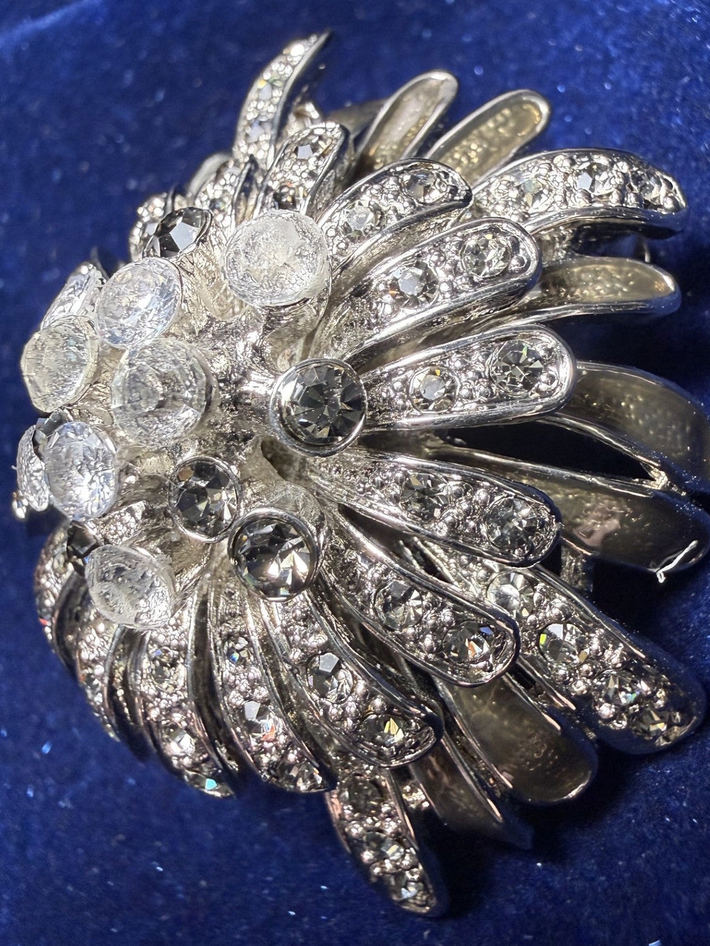 A silver-toned brooch featuring a fan or petal-like openwork design, heavily set with clear rhinestones and accented by larger clear round stones, displayed on a textured dark blue velvet background.