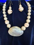 A beaded necklace and matching drop earrings displayed on a dark blue velvet surface. The jewelry features cream-colored round beads, one larger striped bead, and a large iridescent shell pendant on the necklace.