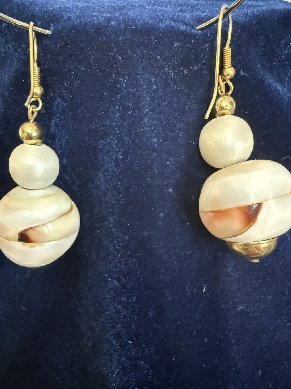 A pair of handmade dangle earrings featuring natural cowrie shells and gold-tone fishhook wires, displayed on a dark blue velvet surface.