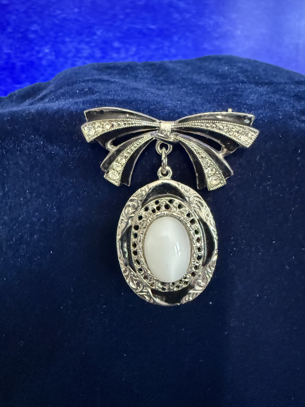 A vintage-style silver bow brooch with a white oval cabochon stone, displayed on a dark blue velvet surface.