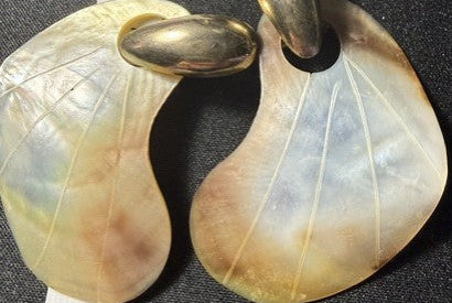 A pair of mother-of-pearl crescent-shaped earrings with gold-toned metal loops, displayed next to a metal ruler on a black background.