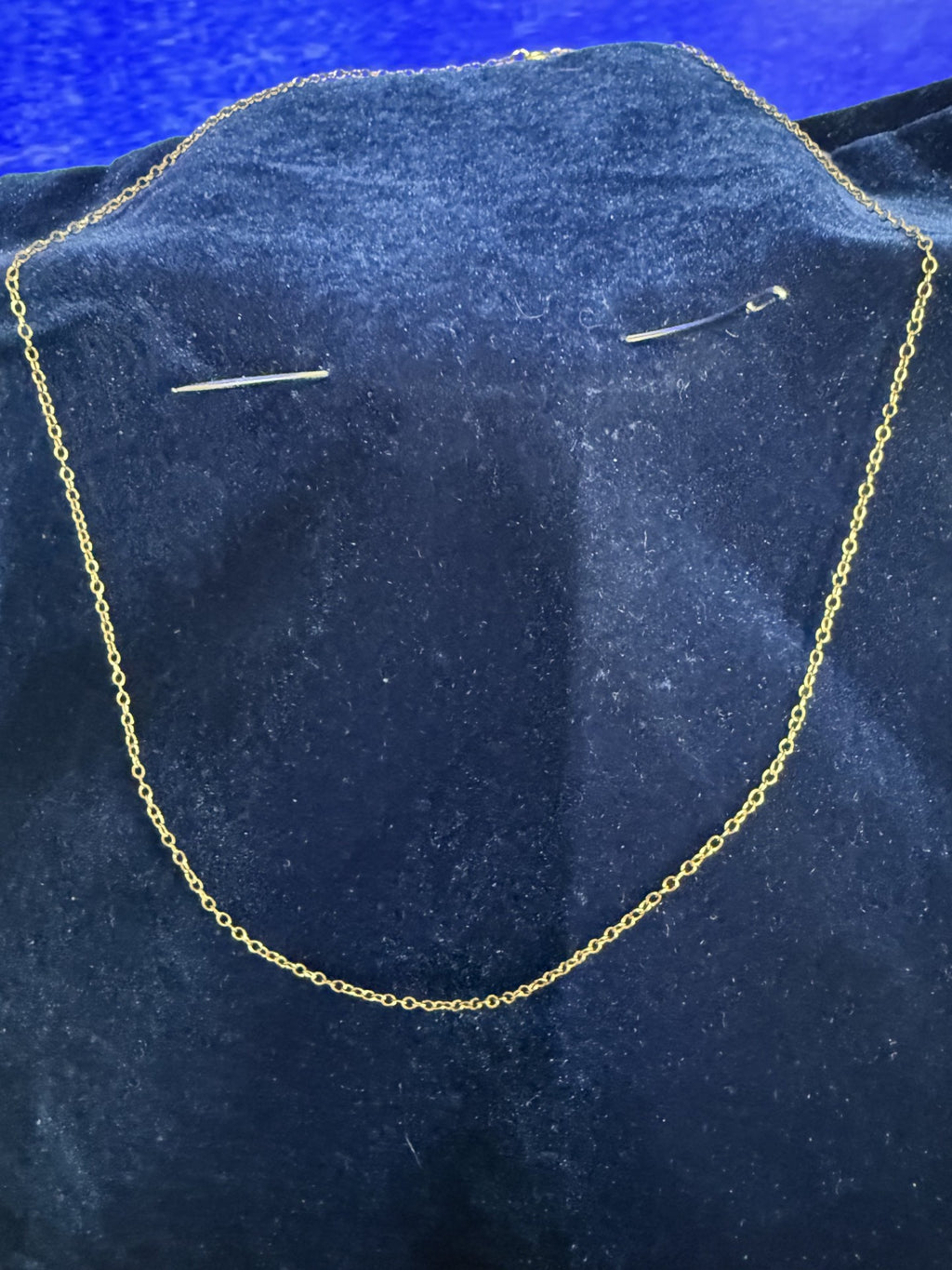 A delicate gold chain necklace displayed on a dark blue velvet jewelry bust, with two small safety pins placed on the bust to hold the chain upright.