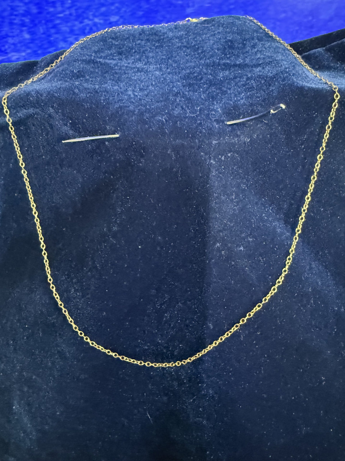 A delicate gold chain necklace displayed on a dark blue velvet jewelry bust, with two small safety pins placed on the bust to hold the chain upright.