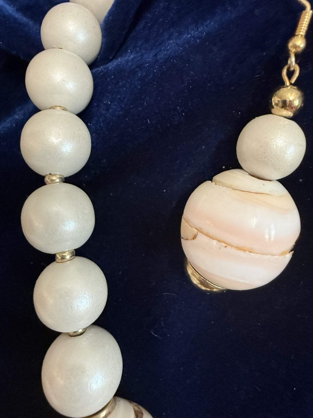 A close-up of a jewelry set featuring a strand of large, glossy white beads with small gold spacers and a matching drop earring with a large white and pale pink stone bead and gold cap, displayed on a dark blue velvet background.