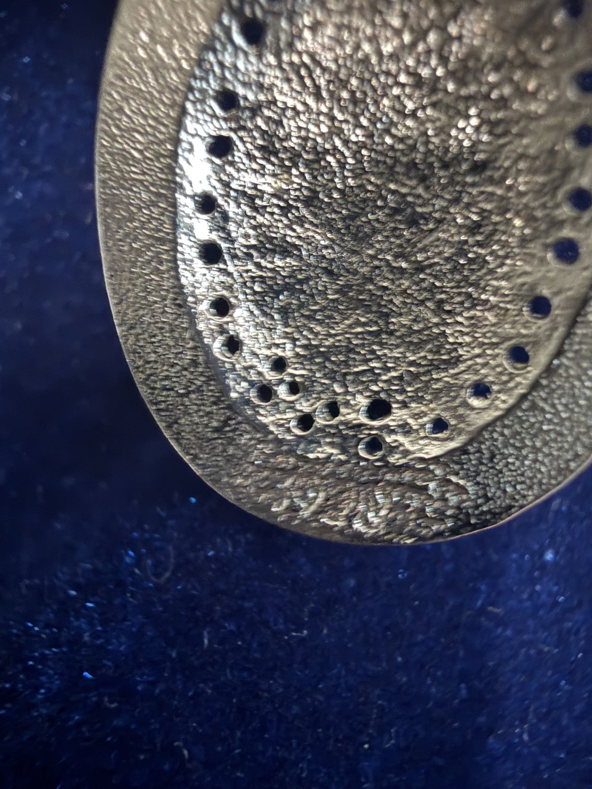 A close-up of a textured metallic circular component with a raised border and small perforations, photographed against a dark blue background.