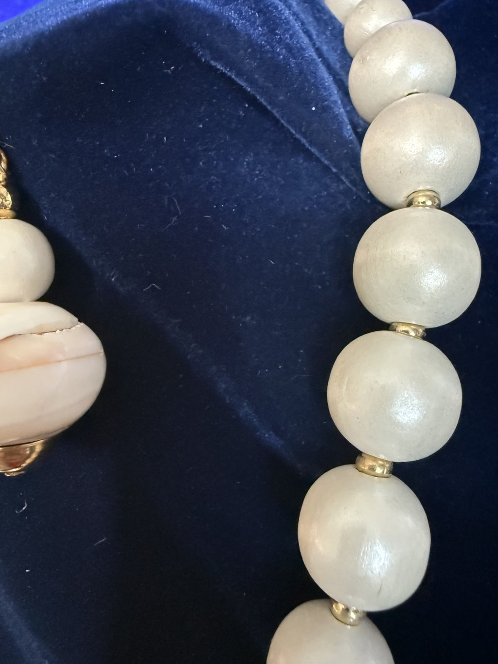 A close-up of a beaded necklace with large, round, off-white beads separated by small gold spacer rings, displayed on a dark blue velvet surface.
