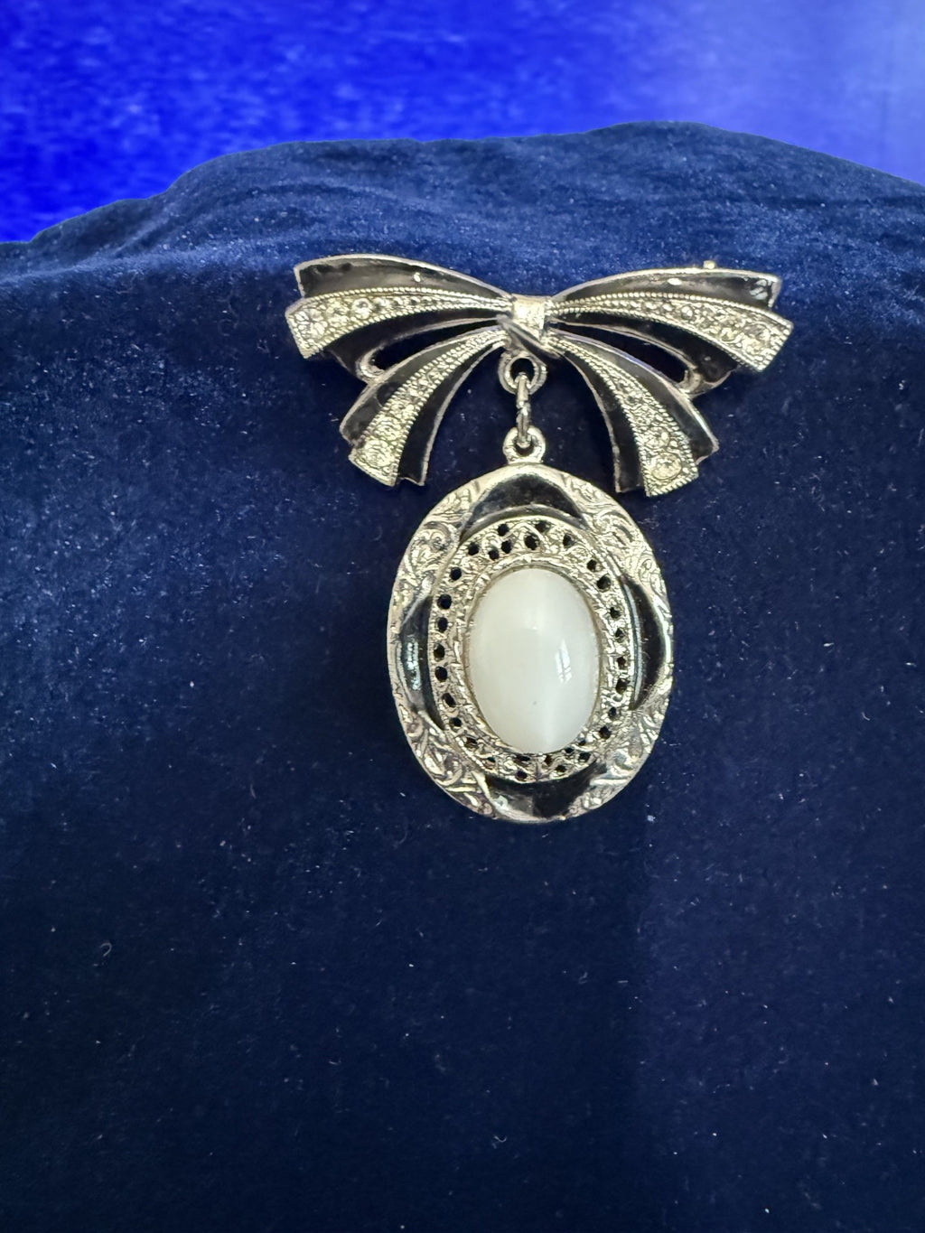 A vintage-style silver-tone brooch shaped like a bow with a dangling oval pendant featuring a milky white cabochon stone, displayed on a dark blue velvet surface.