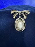 A vintage-style silver-tone brooch shaped like a bow with a dangling oval pendant featuring a milky white cabochon stone, displayed on a dark blue velvet surface.