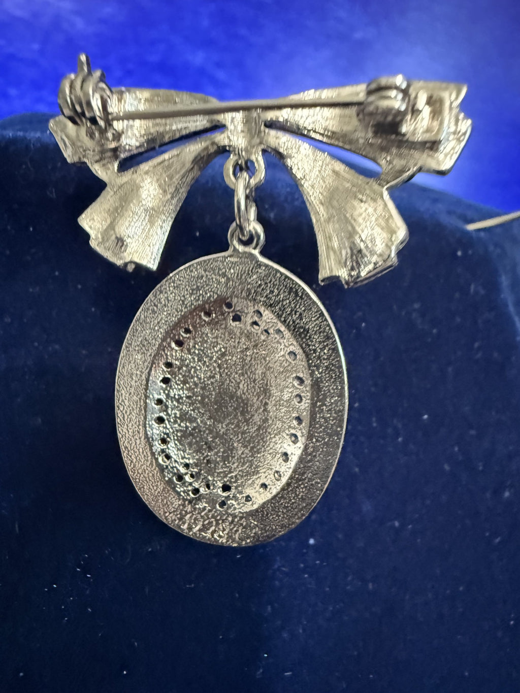 A silver-toned textured brooch featuring a bow-shaped top and a dangling oval pendant, displayed on a dark blue velvet surface.