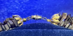 A close-up of a beaded necklace or bracelet with dark brown and gold-toned beads, displayed on a dark blue velvet jewelry stand against a blue background.