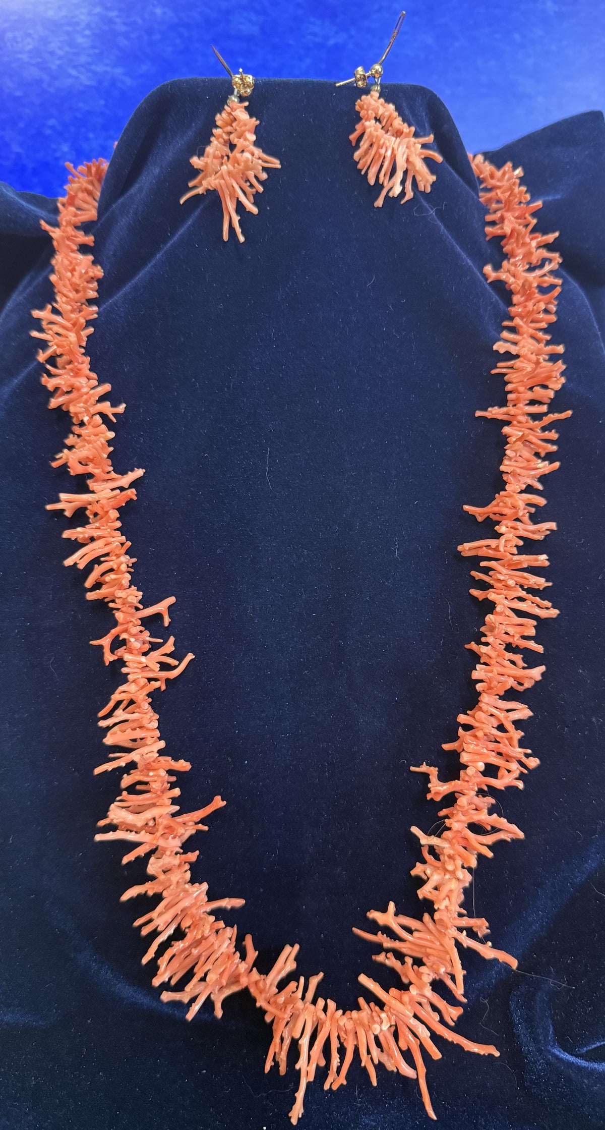 An orange coral necklace and matching drop earrings displayed on a dark blue velvet jewelry bust against a blue background.