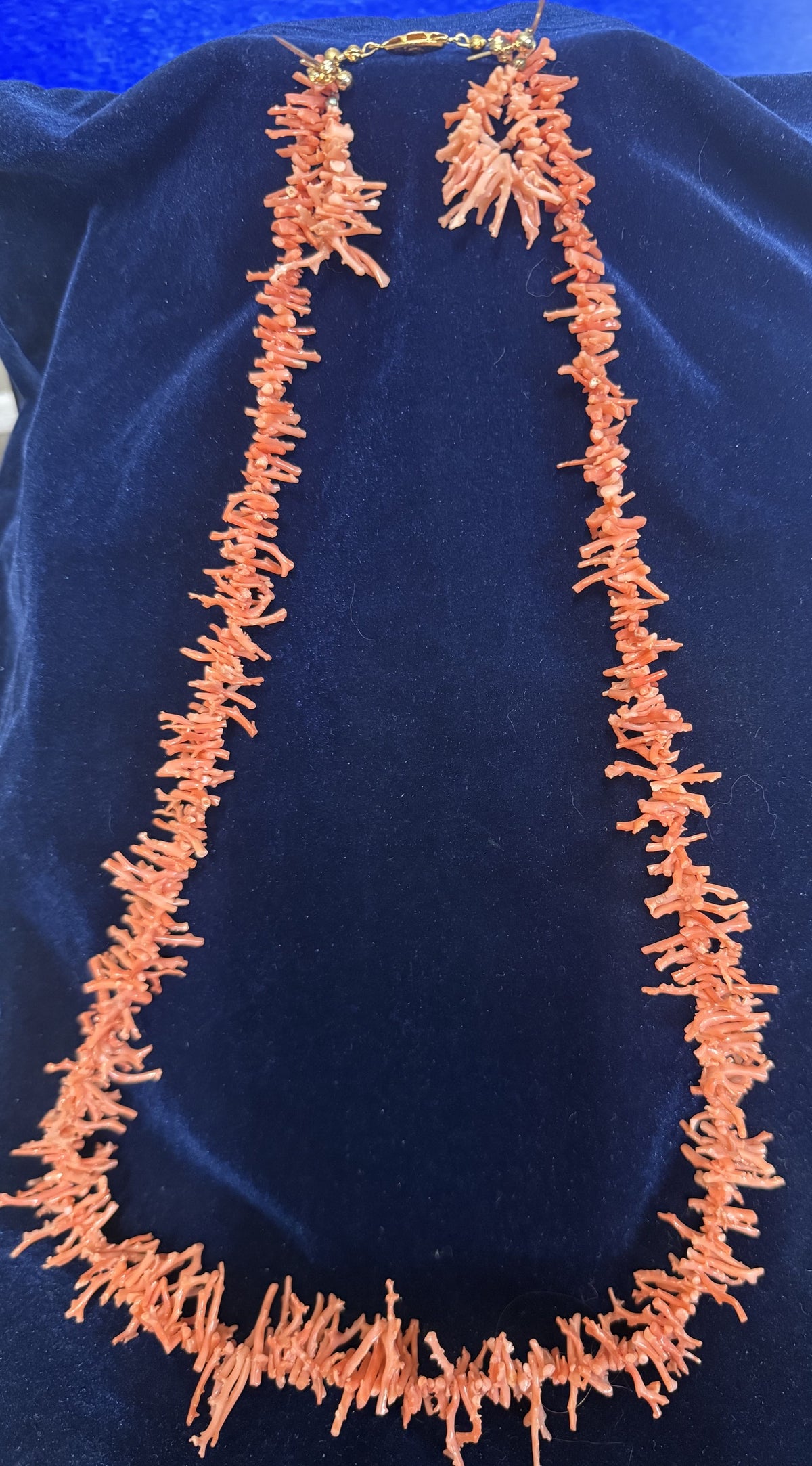 A coral necklace and matching earrings displayed on a dark blue velvet jewelry bust. The coral is a warm orange-pink color, and the clasp is gold-toned.
