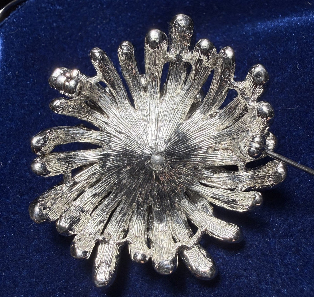 A textured silver-tone flower or sunburst brooch with a central pearl-like accent, displayed on a deep blue velvet surface.