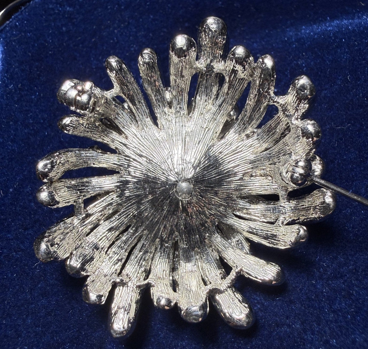 A textured silver-tone flower or sunburst brooch with a central pearl-like accent, displayed on a deep blue velvet surface.