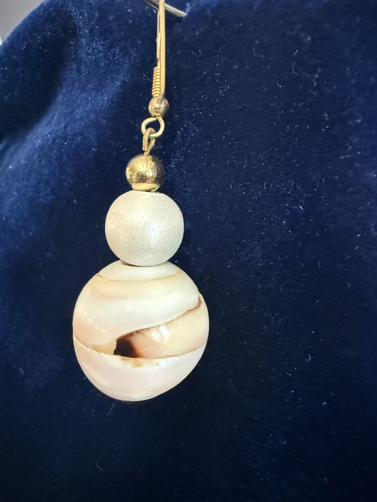 A single dangling earring with a gold-tone hook and beads, featuring a large oval shell bead with cream, tan, and brown marbling above a smaller matte off-white bead, displayed on a dark blue velvet surface.