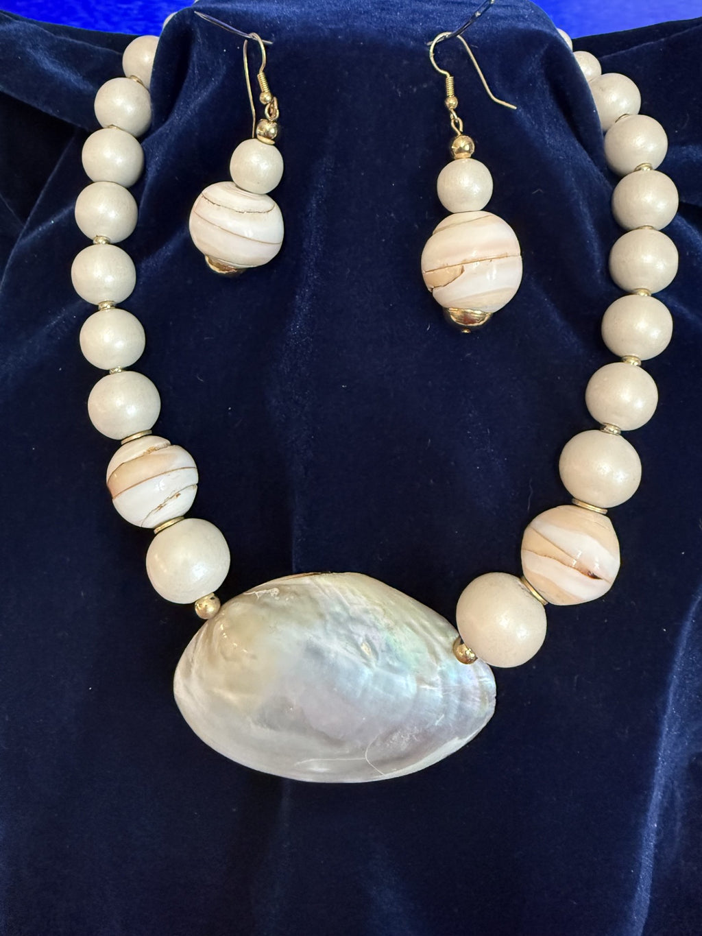A beaded necklace and matching drop earrings displayed on a dark blue velvet surface. The jewelry features cream-colored beads with natural wood striping, separated by small gold spacer beads, with a large iridescent shell pendant on the necklace.