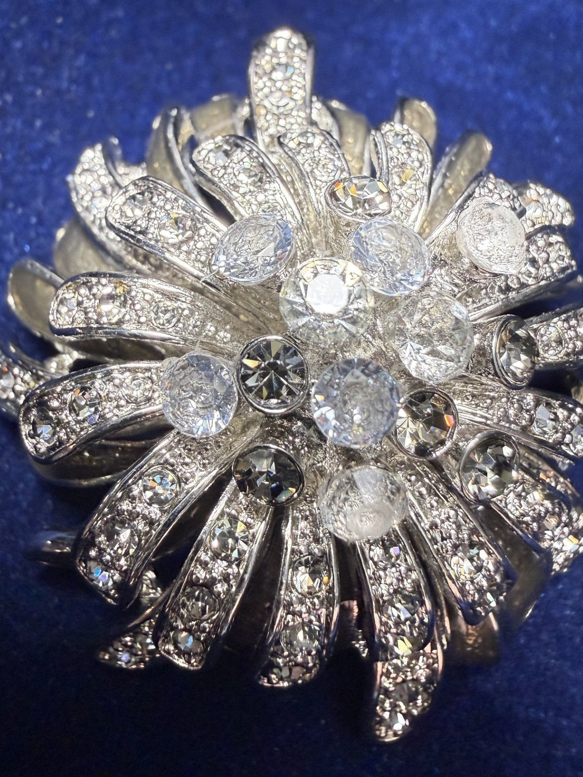 A silver-toned floral brooch or pin embellished with clear rhinestones, displayed on a textured dark blue velvet background.