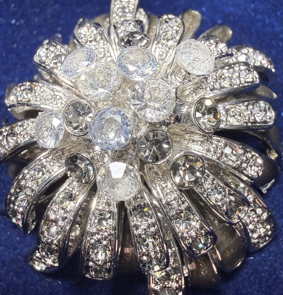 A vintage silver-toned brooch featuring a radiant sunburst design set with clear round and faceted gemstones, displayed on a textured dark blue velvet background.