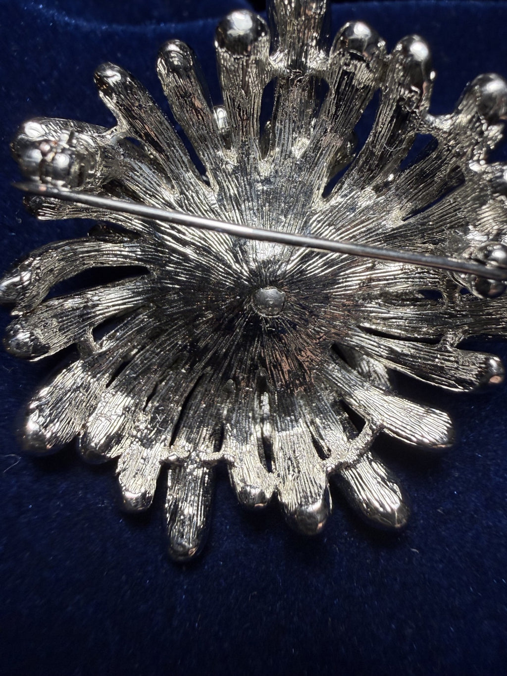 A silver-toned textured brooch with a sunburst or flower-like openwork design, photographed close up on a dark blue velvet surface.