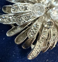 Silver-toned brooch featuring a fan-like arrangement of leaf-shaped metal elements set with clear rhinestones and round clear crystal beads, shown on a dark blue textured background.
