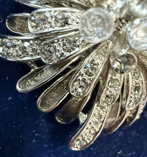 Silver-toned brooch featuring a fan-like arrangement of leaf-shaped metal elements set with clear rhinestones and round clear crystal beads, shown on a dark blue textured background.